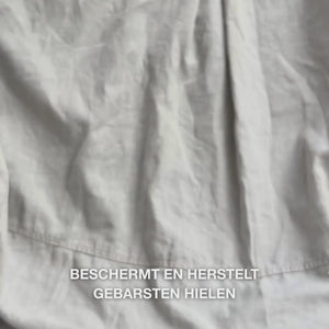 Close-up van een hand die een rijke, romige textuur van Heel Repair in de handpalm neemt en deze zachtjes op droge, gebarsten hielhuid masseert: de dikke substantie wordt zichtbaar opgenomen, verzacht en hydrateert ruwe huid, terwijl 25 % ureum, sheaboter en tea tree olie verzachtend en beschermend werken.
